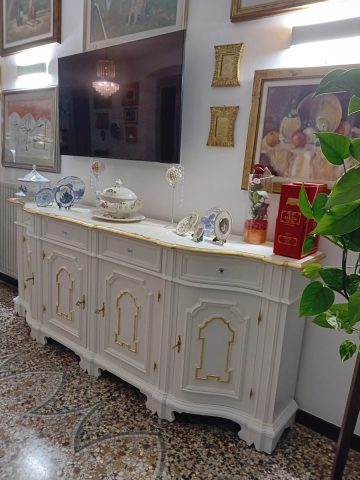 La credenza finita in un bel bianco caldo con riprese a foglia oro sia nelle modanature dei pannelli delle antine sia nel bordo superiore del piano. La credenza finita in un bel bianco caldo con riprese a foglia oro sia nelle modanature dei pannelli delle antine sia nel bordo superiore del piano.